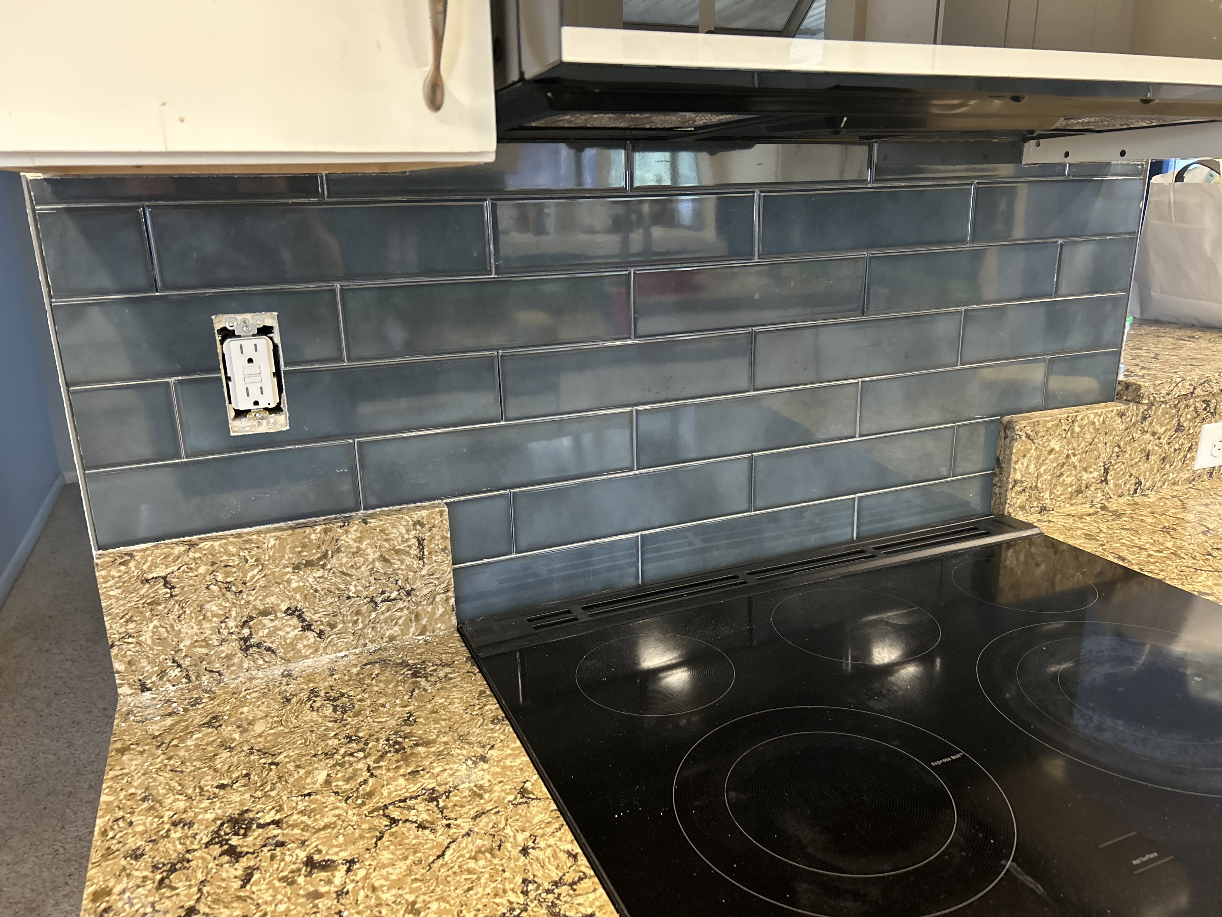 Blue-gray glass subway tile backsplash above a speckled stone countertop beside a black glass cooktop