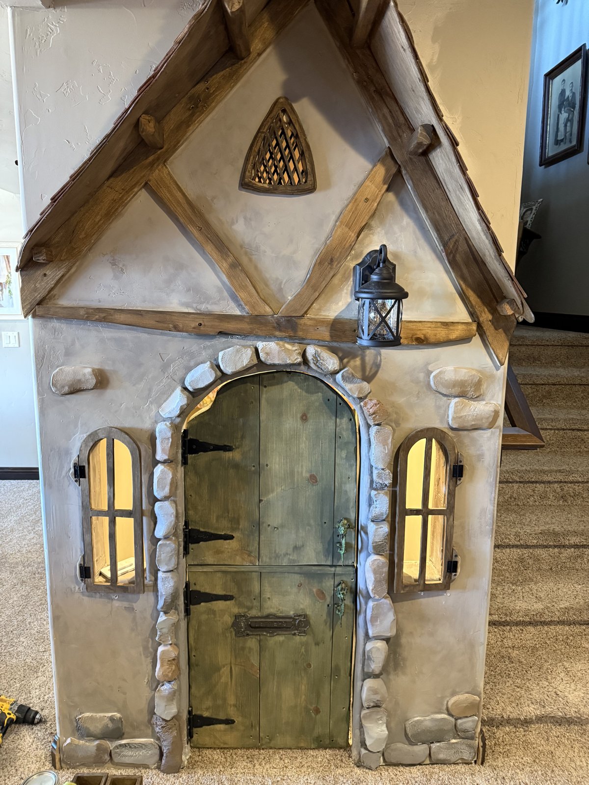 Storybook cottage playhouse built under a staircase, with a faux stone arch, timber gable, arched windows, Dutch door, and a working lantern